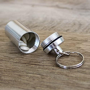 The metal capsule pill box is shown opened, revealing several white tablets inside, with the attached keychain and screw-on lid visible in the foreground. The surface beneath is wooden, providing a natural backdrop.