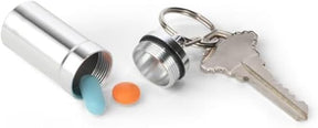 A silver metal capsule pill box is shown partially open with a blue and an orange tablet beside it, along with a key attached to the keychain.