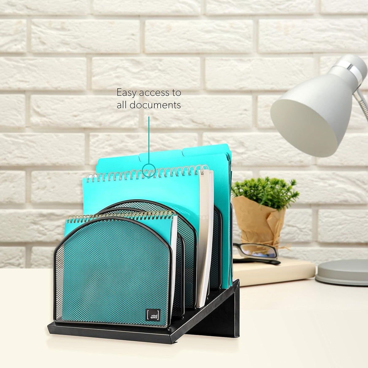 Inclined file sorter in black displaying five compartments filled with various turquoise notebooks and folders, set against a light brick background with a desk lamp and a small green plant nearby.
