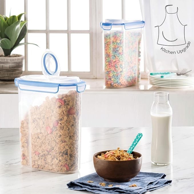 Clear cereal container filled with granola and fruit, featuring a blue lid and a handle, positioned next to a brown bowl of cereal topped with granola on a blue cloth. A glass bottle of milk and colorful cereal in another container are visible in the background.