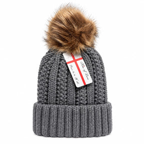 Cozy gray beanie hat with a chunky knit texture, featuring a fluffy brown pom pom on top and a ribbed cuff. A tag is attached, hinting at the brand name.