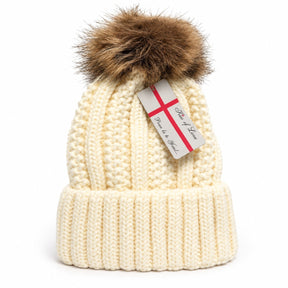 Cream-colored knitted beanie hat featuring a fluffy brown pom pom on top and a ribbed cuff at the base. A tag attached to the hat includes a decorative design.