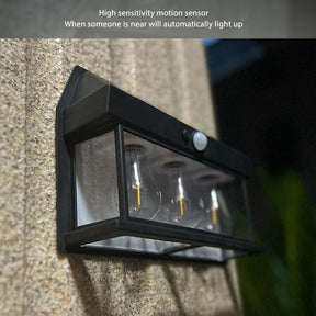 The outdoor light features a sleek black frame with three glass bulbs inside, mounted on a textured wall. A motion sensor is positioned at the top, enabling automatic lighting when someone approaches.