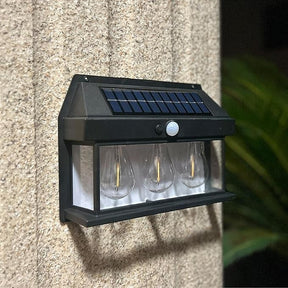 Three clear bulb-shaped lights are housed in a black frame with a solar panel and motion sensor on top, mounted against a textured wall. The design features a sleek, modern aesthetic suitable for outdoor use.