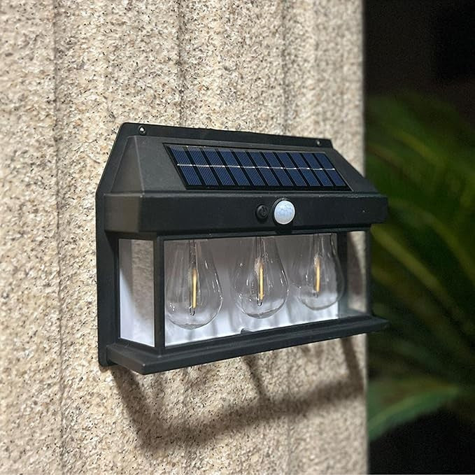 Three clear bulb-shaped lights are housed in a black frame with a solar panel and motion sensor on top, mounted against a textured wall. The design features a sleek, modern aesthetic suitable for outdoor use.