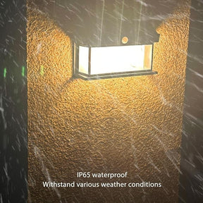 The solar motion sensor light is mounted on a textured wall, emitting a bright light against a backdrop of falling rain, highlighting its IP65 waterproof capability. The light features a sleek, rectangular design with clear sides and a sturdy metal frame.