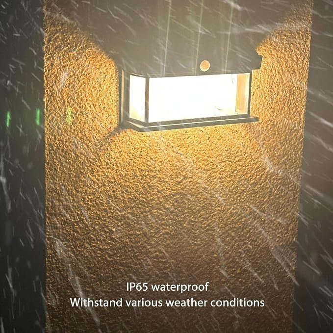 The solar motion sensor light is mounted on a textured wall, emitting a bright light against a backdrop of falling rain, highlighting its IP65 waterproof capability. The light features a sleek, rectangular design with clear sides and a sturdy metal frame.
