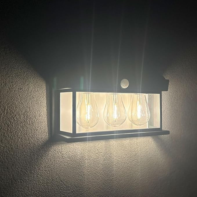 The solar motion sensor light features a sleek black frame with three illuminated bulb-like fixtures, mounted against a textured wall. A motion sensor is visible at the top, enhancing its modern design.