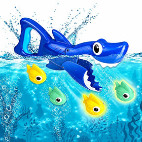 Bright blue shark-shaped water toy partially submerged in water, with three colorful fish (two yellow and one green) nearby, surrounded by bubbles and aquatic plants.