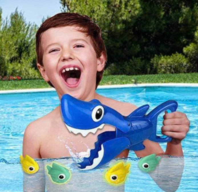 A child joyfully smiles while holding a blue plastic shark toy in a swimming pool, with colorful fish toys floating in the water around him. The background features lush greenery and the edge of the pool.