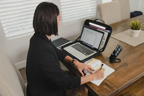 A person in a black blazer is seated at a wooden table, using a laptop open inside the portable briefcase while writing in a notebook. A smartphone is positioned on a stand next to them, and there is a potted plant visible in the background.