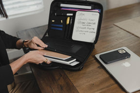 A person is organizing notebooks and a whiteboard tablet inside the open briefcase, which features multiple compartments for office supplies. A laptop and a smartphone are visible on the wooden table next to the briefcase.