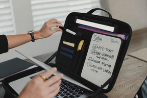 A hand reaches for a pen on the interior of a black briefcase, which features a whiteboard section with handwritten notes and checklists, alongside colorful sticky notes and pens. A silver laptop is positioned in front of the briefcase, set on a wooden table.
